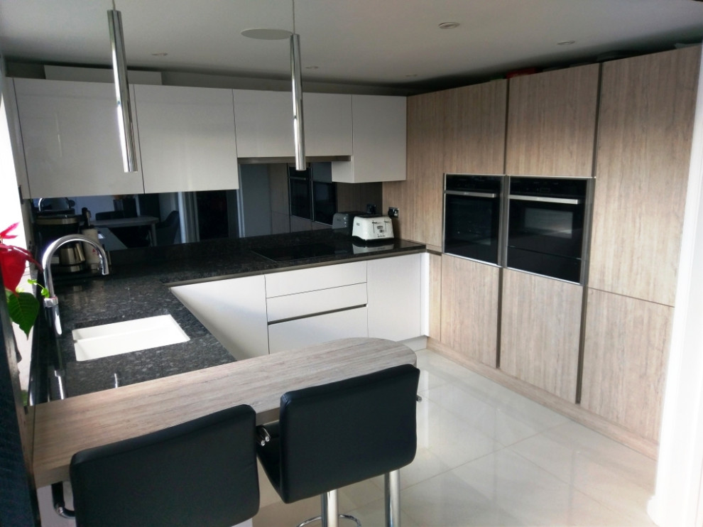 This is an example of a small modern u-shaped eat-in kitchen in London with an integrated sink, flat-panel cabinets, granite benchtops, metallic splashback, glass sheet splashback, stainless steel appliances, ceramic floors, a peninsula, beige floor and black benchtop.