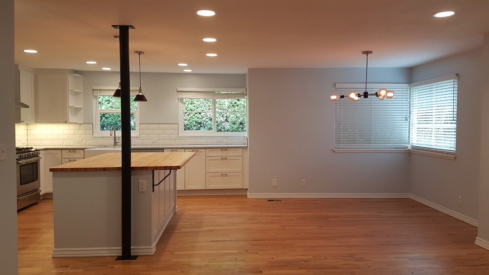 Photo of a mid-sized contemporary l-shaped open plan kitchen in Seattle with a farmhouse sink, recessed-panel cabinets, white cabinets, marble benchtops, white splashback, ceramic splashback, stainless steel appliances, medium hardwood floors, with island, brown floor and grey benchtop.