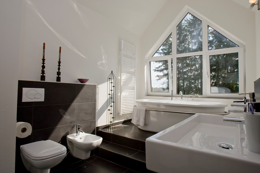 This is an example of a mid-sized contemporary bathroom in Other with a freestanding tub, a wall-mount toilet, gray tile, white walls and a wall-mount sink.
