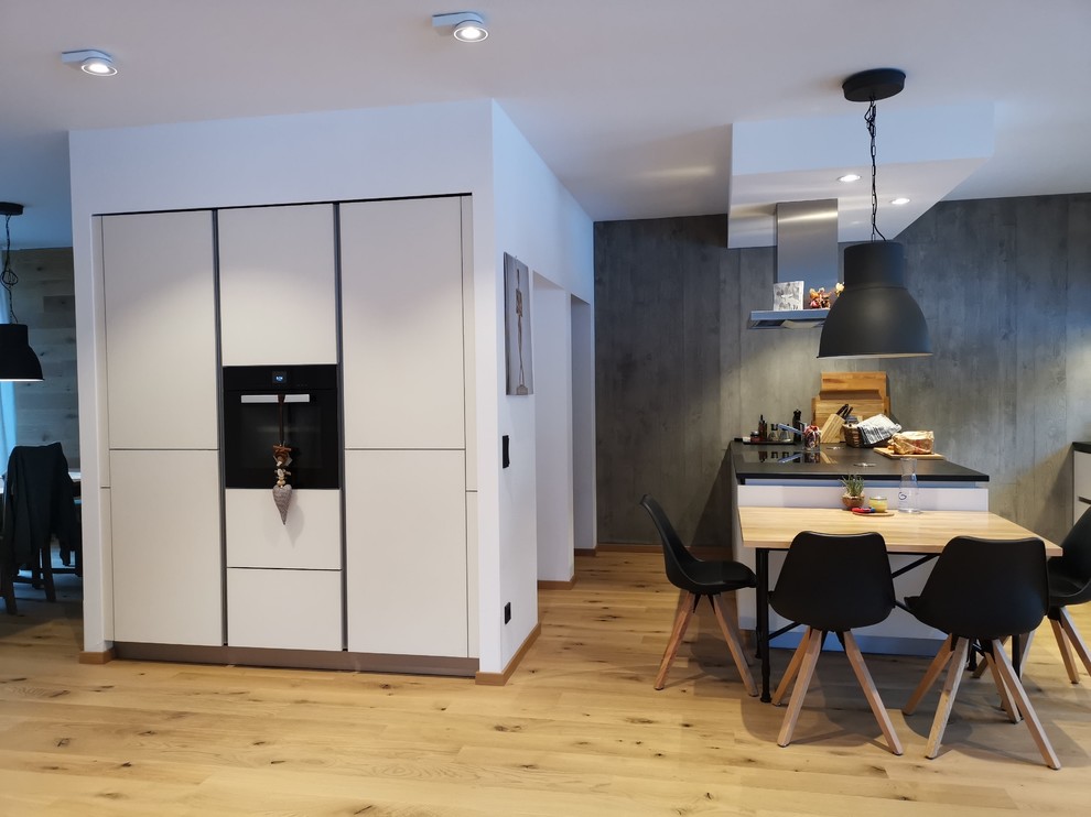 Photo of a mid-sized contemporary galley open plan kitchen in Dresden with flat-panel cabinets, white cabinets, granite benchtops, grey splashback, black appliances, light hardwood floors, a peninsula and black benchtop.