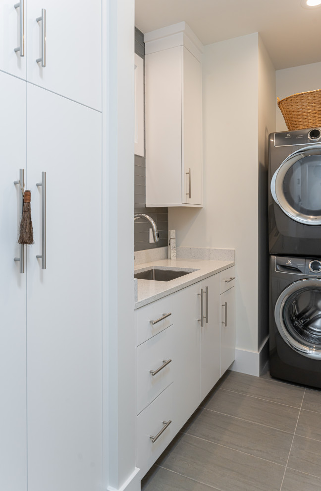 Small modern galley dedicated laundry room in Other with an undermount sink, flat-panel cabinets, white cabinets, quartz benchtops, grey splashback, porcelain splashback, white walls, porcelain floors, a stacked washer and dryer, grey floor and white benchtop.