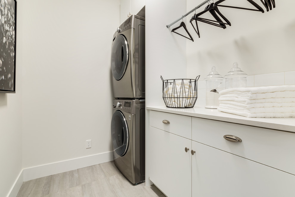 Inspiration for a mid-sized contemporary single-wall dedicated laundry room in Calgary with flat-panel cabinets, white cabinets, solid surface benchtops, white walls, porcelain floors, a stacked washer and dryer, grey floor and white benchtop.