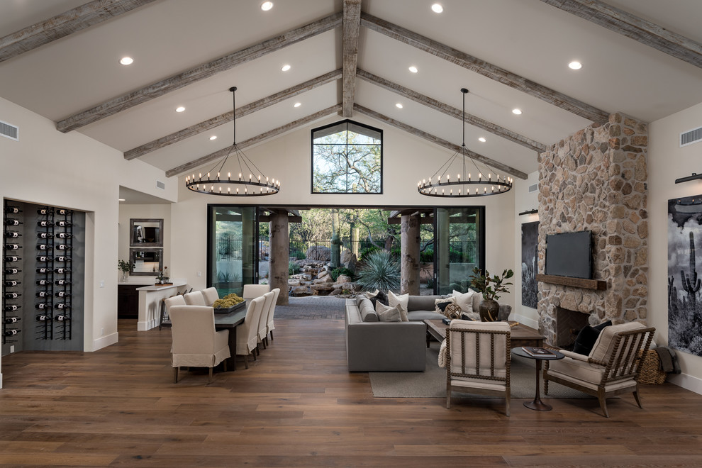 This is an example of an open concept family room in Phoenix with a home bar, beige walls, a standard fireplace, a stone fireplace surround, a wall-mounted tv, brown floor and medium hardwood floors.