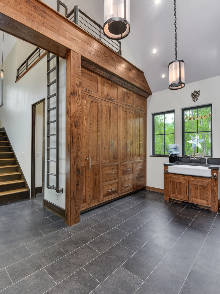 This is an example of a large transitional u-shaped utility room in Other with a farmhouse sink, shaker cabinets, medium wood cabinets, porcelain floors, a side-by-side washer and dryer, grey floor and grey walls.