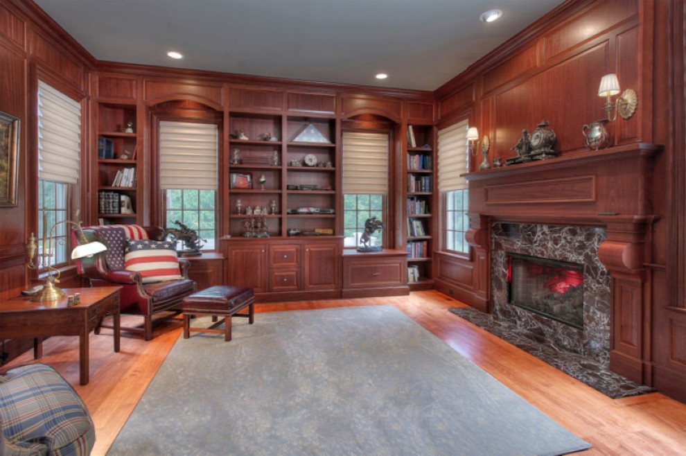 This is an example of a large traditional home office in New York with a standard fireplace, a library, brown walls, medium hardwood floors, a wood fireplace surround, a freestanding desk and brown floor.