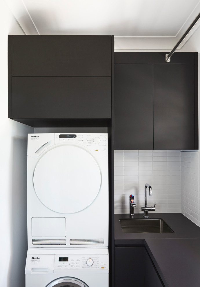 Inspiration for a small contemporary l-shaped utility room in Sydney with flat-panel cabinets, black cabinets, grey floors, white splashback, cement tile splashback, white walls, ceramic flooring, a stacked washer and dryer, a submerged sink and black worktops.