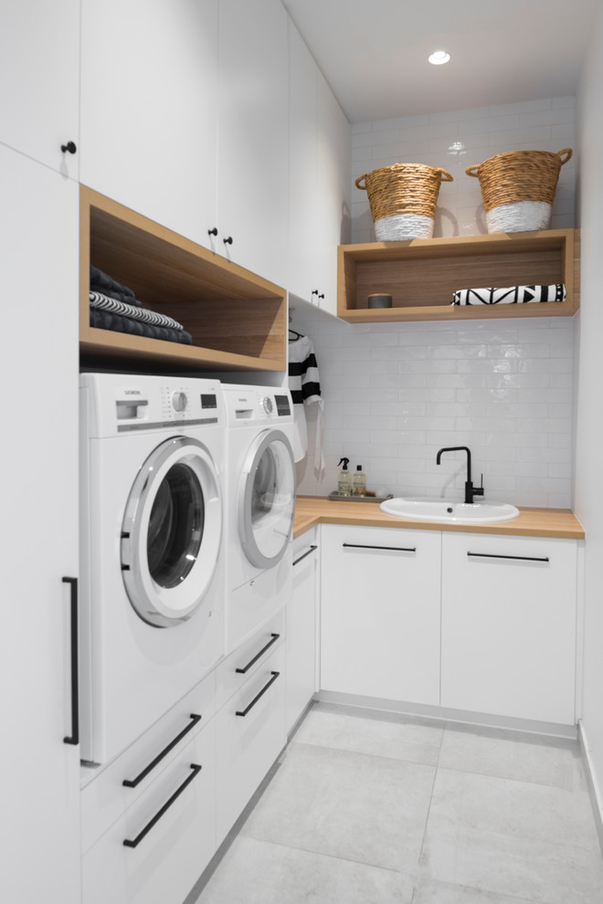 Photo of a small beach style l-shaped dedicated laundry room in Adelaide with a drop-in sink, white cabinets, wood benchtops, white walls, ceramic floors, a side-by-side washer and dryer, brown benchtop, flat-panel cabinets and grey floor.