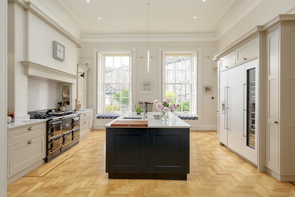 This is an example of a large transitional galley eat-in kitchen in Edinburgh with an integrated sink, shaker cabinets, blue cabinets, quartz benchtops, panelled appliances, light hardwood floors, with island, brown floor, grey benchtop, grey splashback and marble splashback.