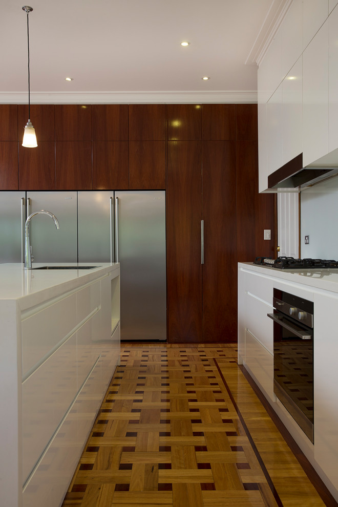 Design ideas for a contemporary l-shaped kitchen in Sydney with white cabinets, quartz benchtops, white splashback, stainless steel appliances, with island, white benchtop, an undermount sink, flat-panel cabinets, glass sheet splashback, medium hardwood floors and brown floor.