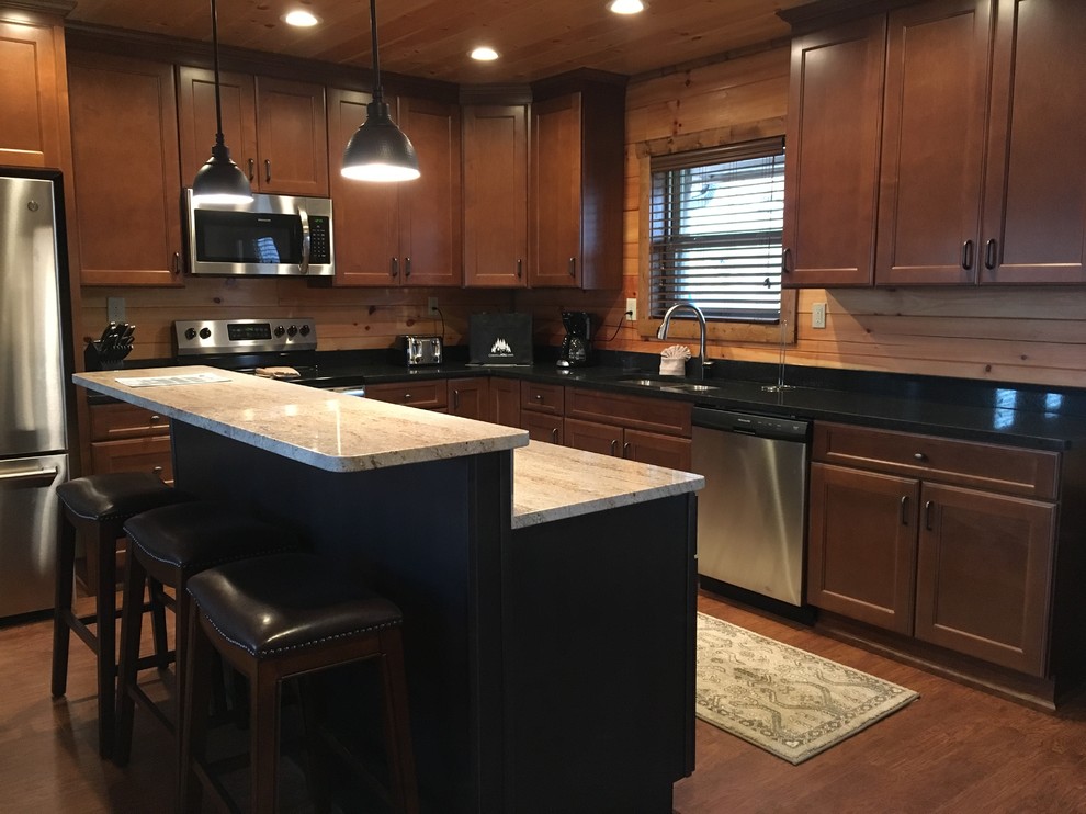 Mountain style l-shaped laminate floor and brown floor eat-in kitchen photo with a double-bowl sink, flat-panel cabinets, dark wood cabinets, granite countertops, black backsplash, stainless steel appliances, an island and black countertops
