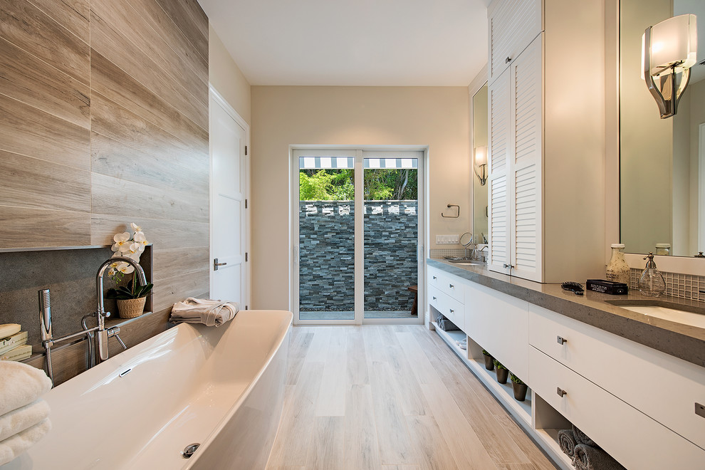 This is an example of an expansive contemporary master bathroom in Miami with flat-panel cabinets, white cabinets, a freestanding tub, a corner shower, a one-piece toilet, gray tile, glass tile, beige walls, light hardwood floors, a drop-in sink, limestone benchtops, beige floor and a hinged shower door.