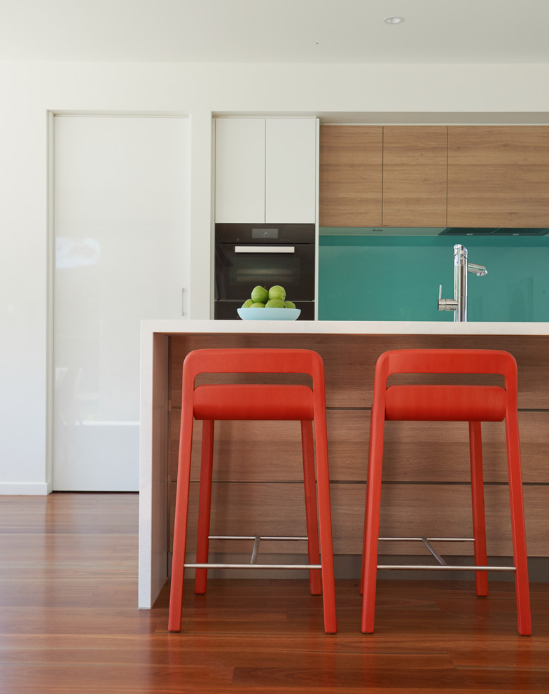 Photo of a mid-sized contemporary galley kitchen in Melbourne with flat-panel cabinets, with island, solid surface benchtops, green splashback, glass sheet splashback, black appliances, medium hardwood floors and light wood cabinets.