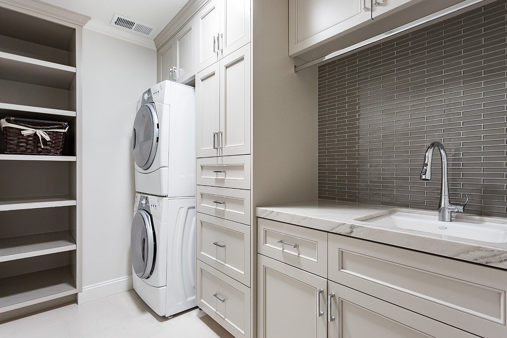 Photo of a transitional single-wall laundry room in San Francisco with an undermount sink, beige cabinets, marble benchtops, grey walls, a stacked washer and dryer, white benchtop and recessed-panel cabinets.