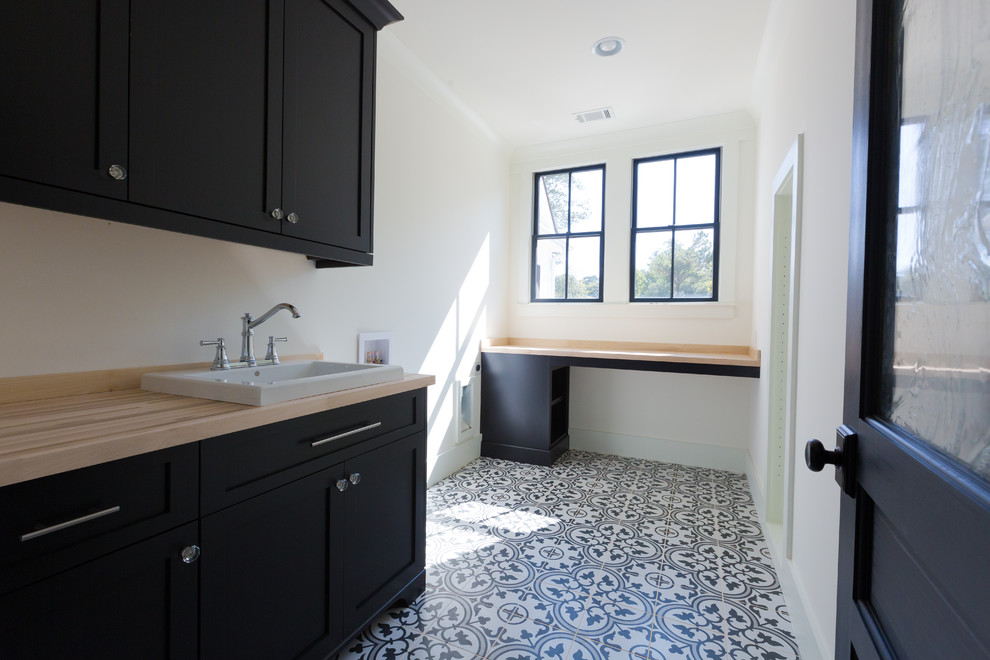 Photo of a mid-sized transitional l-shaped dedicated laundry room in Atlanta with a drop-in sink, shaker cabinets, black cabinets, wood benchtops, white walls, ceramic floors, a side-by-side washer and dryer and multi-coloured floor.