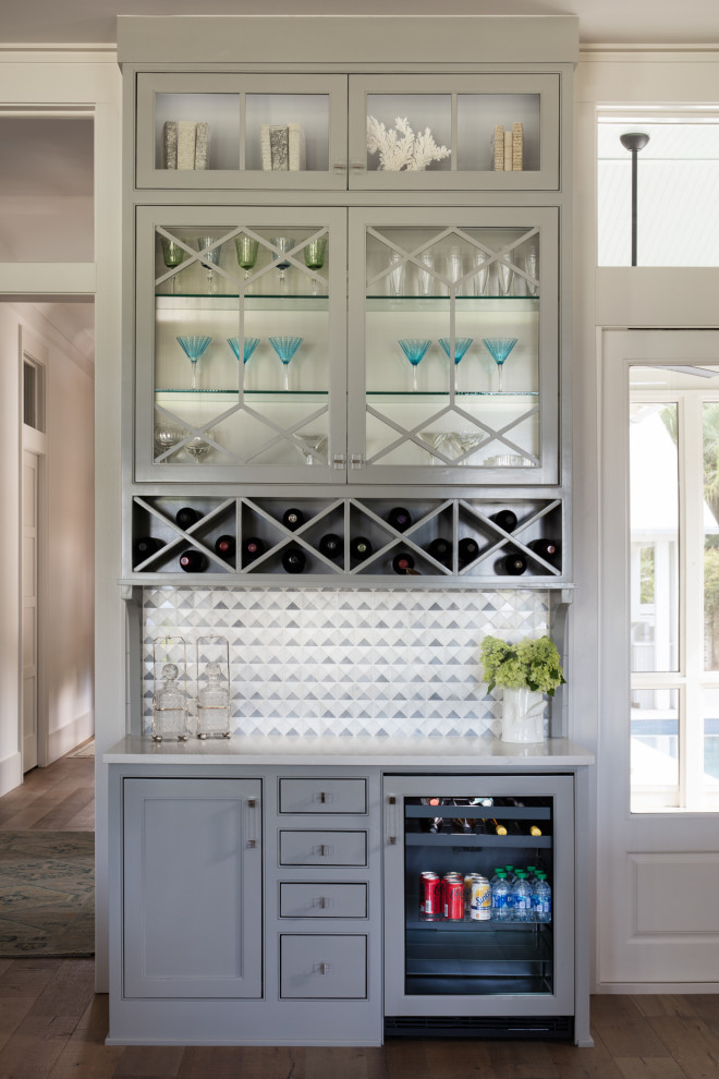 Inspiration for a large beach style home bar in Birmingham with recessed-panel cabinets, grey cabinets, cement tile splashback, medium hardwood floors, brown floor and white benchtop.