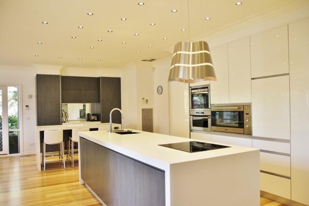 Photo of a large modern l-shaped open plan kitchen in Melbourne with an undermount sink, flat-panel cabinets, white cabinets, quartz benchtops, mirror splashback, stainless steel appliances, light hardwood floors and multiple islands.