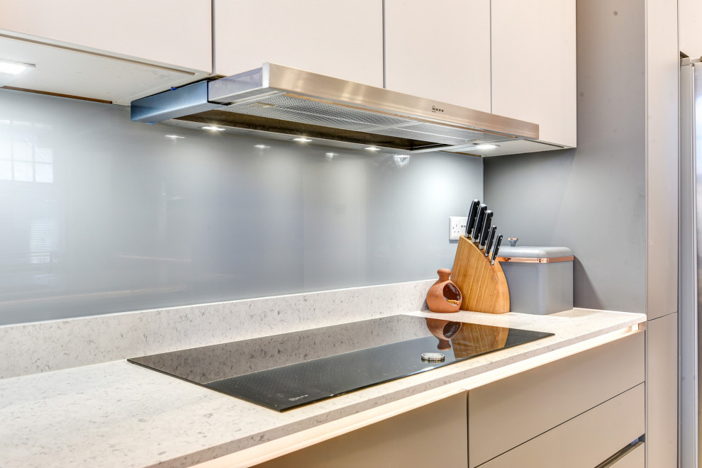 Expansive modern grey and white single-wall kitchen/diner in Sussex with a double-bowl sink, flat-panel cabinets, grey cabinets, quartz worktops, grey splashback, glass sheet splashback, black appliances, vinyl flooring, an island, grey floors, white worktops and feature lighting.