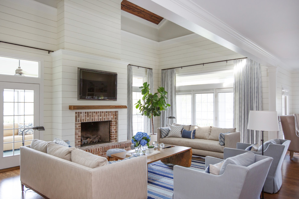 This is an example of a beach style family room in Jacksonville with white walls, medium hardwood floors, a standard fireplace, a brick fireplace surround, a wall-mounted tv and brown floor.