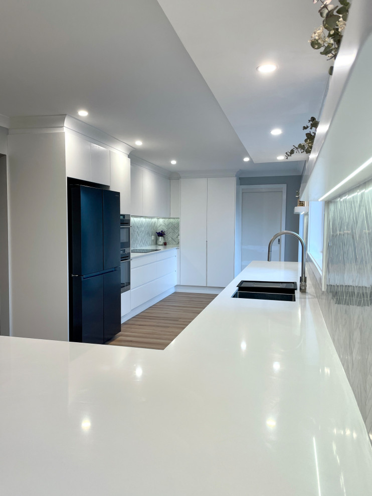 Photo of a large modern galley eat-in kitchen in Sydney with a drop-in sink, flat-panel cabinets, white cabinets, quartz benchtops, grey splashback, marble splashback, black appliances, light hardwood floors, brown floor and white benchtop.