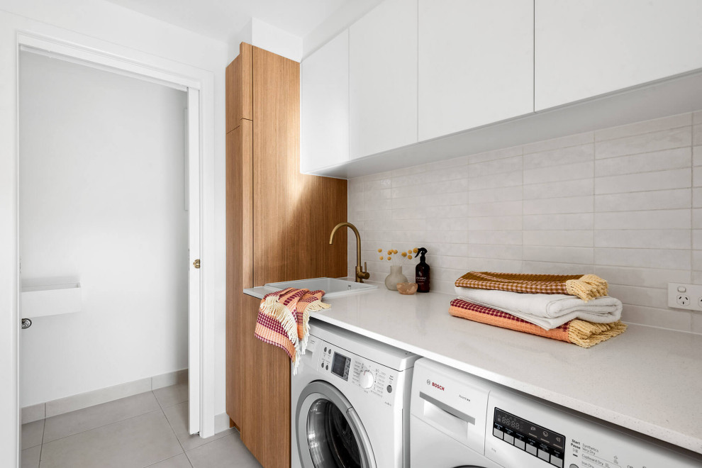 Inspiration for a mid-sized modern single-wall utility room in Melbourne with a drop-in sink, flat-panel cabinets, medium wood cabinets, quartz benchtops, white splashback, ceramic splashback, white walls, ceramic floors, a side-by-side washer and dryer, beige floor and white benchtop.