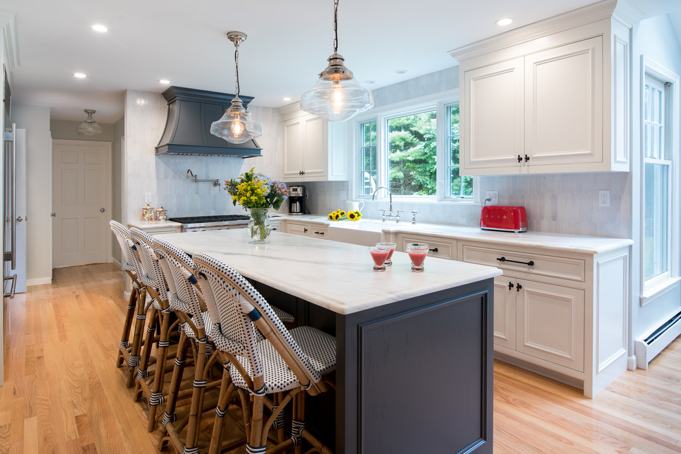 Example of a transitional u-shaped medium tone wood floor and brown floor kitchen design in Boston with a farmhouse sink, beaded inset cabinets, white cabinets, gray backsplash, stainless steel appliances, an island and white countertops