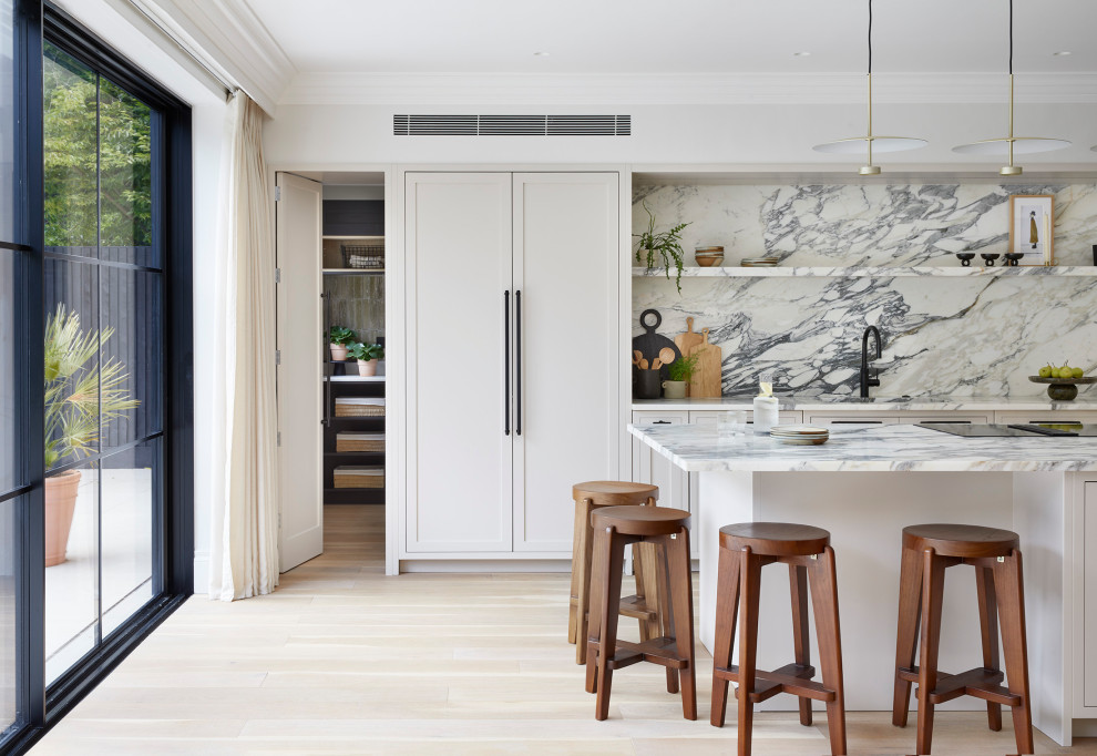 Photo of a medium sized scandinavian cream and black l-shaped open plan kitchen in London with a built-in sink, shaker cabinets, beige cabinets, marble worktops, white splashback, marble splashback, stainless steel appliances, light hardwood flooring, an island, white worktops and feature lighting.