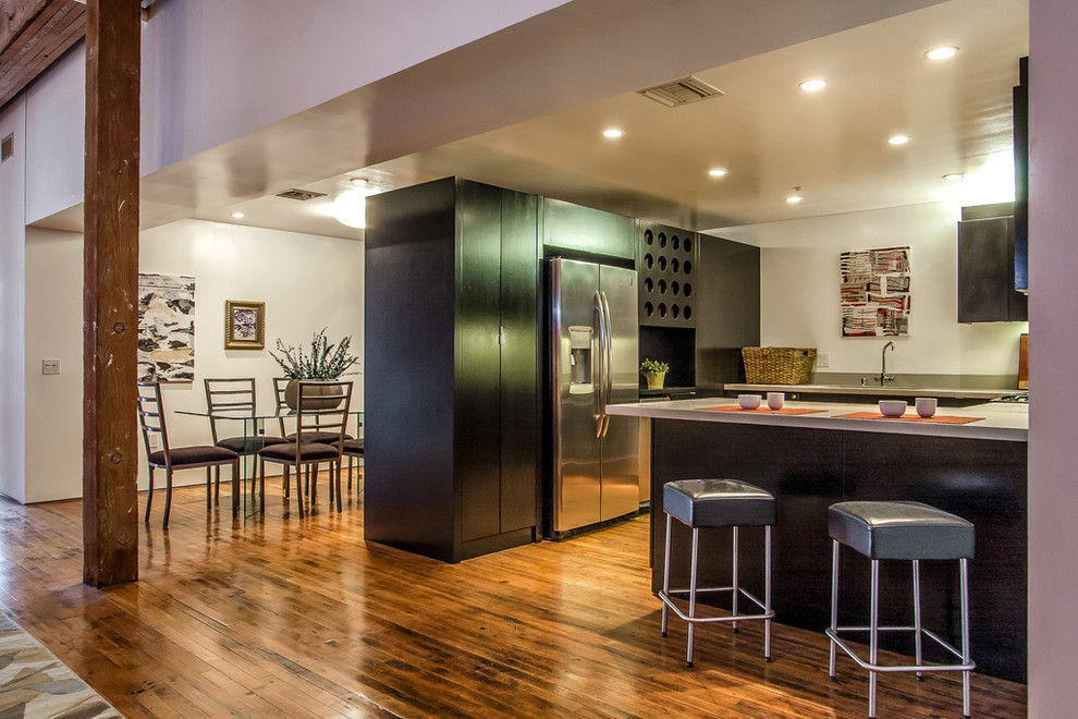 This is an example of a mid-sized industrial u-shaped eat-in kitchen in Los Angeles with flat-panel cabinets, black cabinets, stainless steel appliances, an undermount sink, solid surface benchtops, medium hardwood floors and a peninsula.