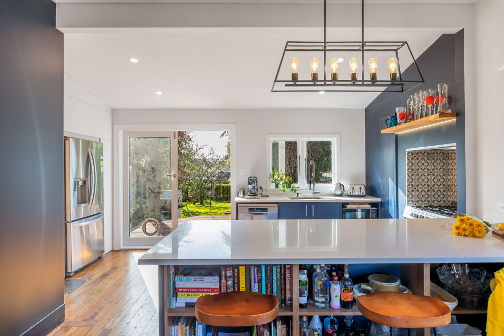 Inspiration for a medium sized eclectic u-shaped kitchen in Auckland with a double-bowl sink, flat-panel cabinets, blue cabinets, engineered stone countertops, grey splashback, ceramic splashback, stainless steel appliances, medium hardwood flooring, brown floors, white worktops and a coffered ceiling.