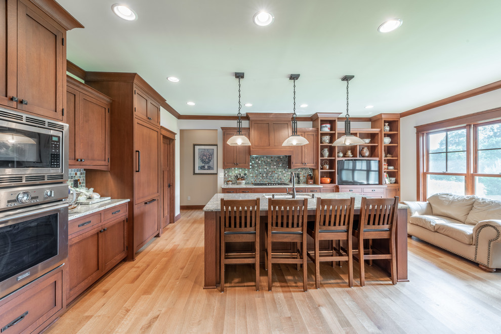 This is an example of a mid-sized arts and crafts eat-in kitchen in Milwaukee with an undermount sink, beaded inset cabinets, medium wood cabinets, granite benchtops, metallic splashback, glass sheet splashback, panelled appliances, light hardwood floors, with island, yellow floor and beige benchtop.