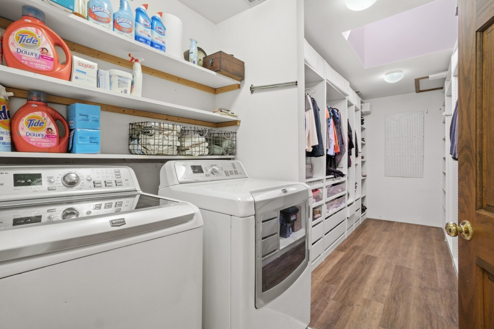 Klassisk inredning av en stor parallell tvättstuga enbart för tvätt, med öppna hyllor, vita skåp, vita väggar, en tvättmaskin och torktumlare bredvid varandra, ljust trägolv och brunt golv