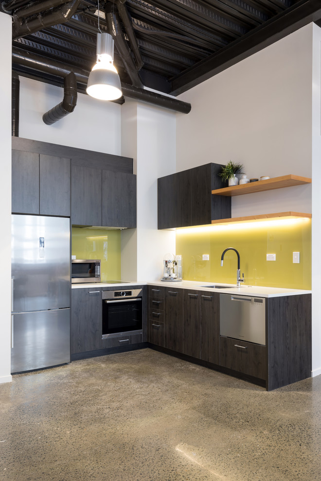 This is an example of an industrial l-shaped kitchen in Auckland with an undermount sink, flat-panel cabinets, grey cabinets, yellow splashback, glass sheet splashback, stainless steel appliances, no island and grey floor.