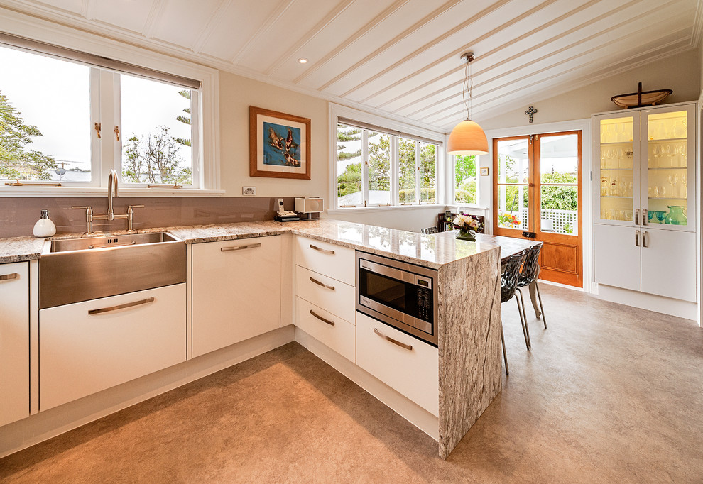 This is an example of a mid-sized traditional u-shaped eat-in kitchen in Auckland with white cabinets, granite benchtops, glass sheet splashback, stainless steel appliances, linoleum floors, a farmhouse sink, flat-panel cabinets, brown splashback, no island and grey floor.