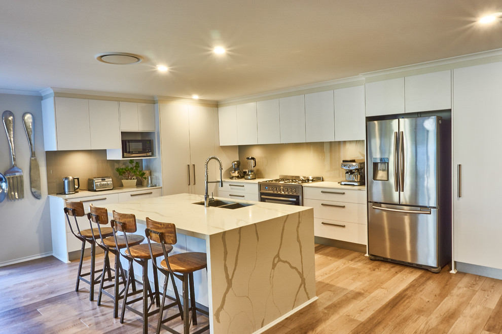 Photo of a mid-sized contemporary l-shaped open plan kitchen in Gold Coast - Tweed with a double-bowl sink, flat-panel cabinets, white cabinets, quartz benchtops, beige splashback, glass sheet splashback, stainless steel appliances, laminate floors, with island, brown floor and beige benchtop.