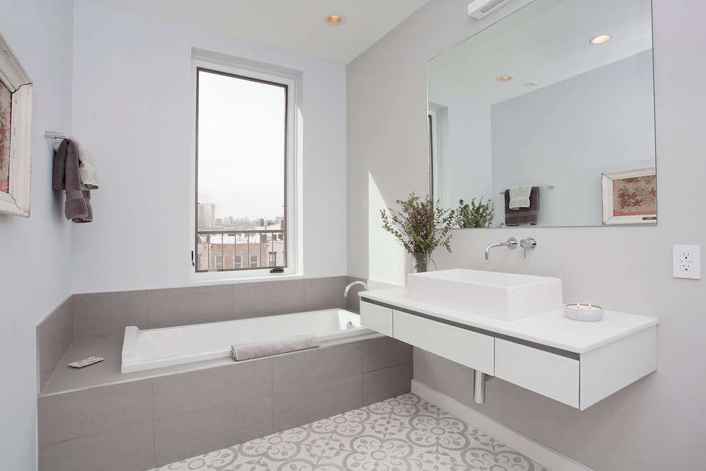 Photo of a mid-sized scandinavian bathroom in New York with a vessel sink, a drop-in tub, gray tile and grey walls.