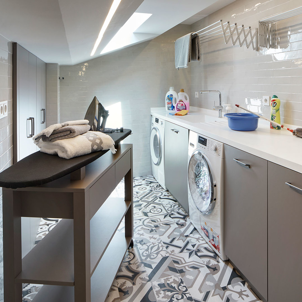 This is an example of a contemporary single-wall dedicated laundry room in Barcelona with an integrated sink, flat-panel cabinets, grey cabinets, white walls, a side-by-side washer and dryer, multi-coloured floor and white benchtop.