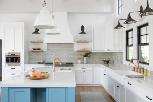 Coastal farmhouse kitchen with white shaker premium RTA cabinets Canada , a blue island with quartz countertop, brass fixtures, gray subway tile backsplash, and black-framed windows.