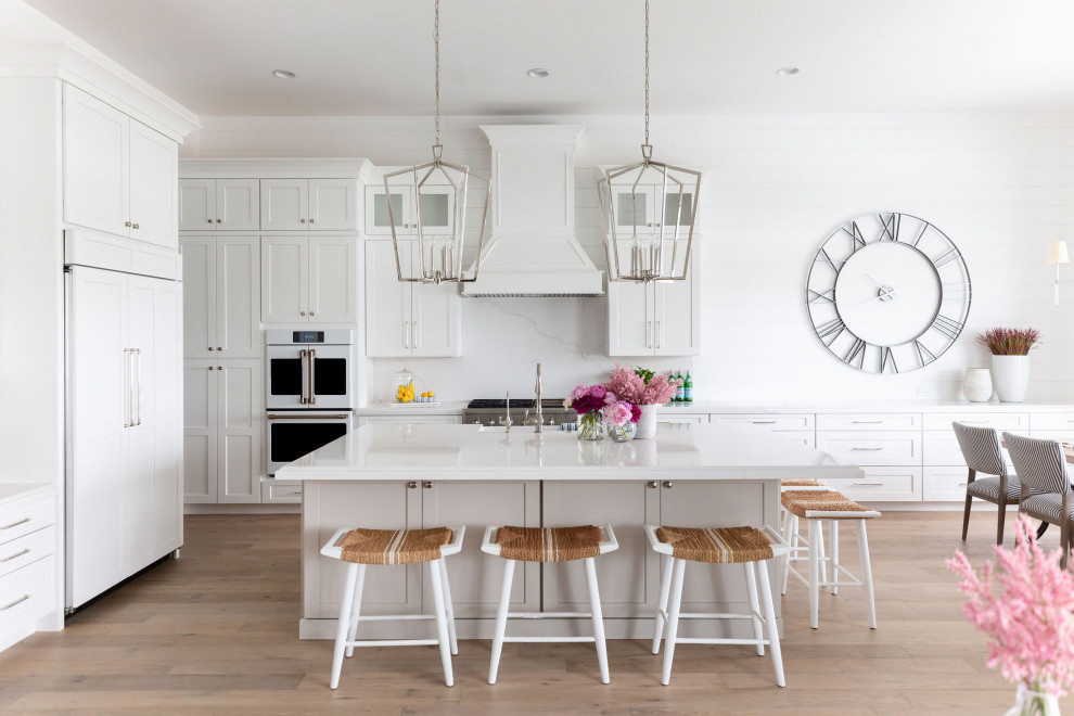 This is an example of a large beach style open plan kitchen in Phoenix with a farmhouse sink, shaker cabinets, white cabinets, quartz benchtops, white splashback, engineered quartz splashback, white appliances, light hardwood floors, with island, beige floor and white benchtop.