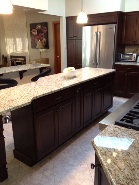 Transitional porcelain tile eat-in kitchen photo with a double-bowl sink, recessed-panel cabinets, brown cabinets, granite countertops, beige backsplash, stone slab backsplash, stainless steel appliances and an island