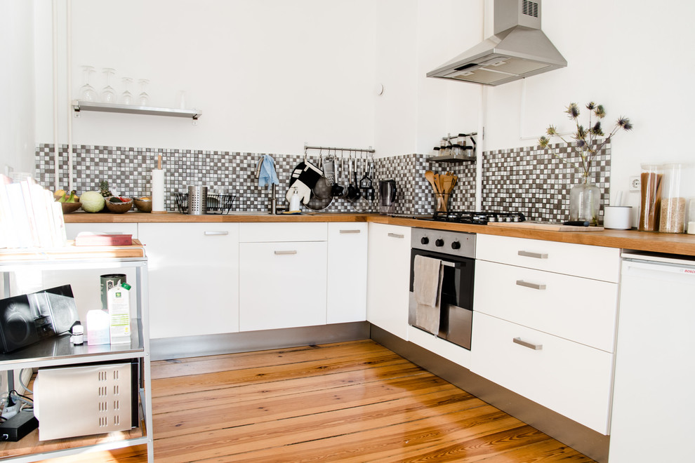 Design ideas for a mid-sized scandinavian separate kitchen in Berlin with a drop-in sink, flat-panel cabinets, white cabinets, wood benchtops, grey splashback, mosaic tile splashback, stainless steel appliances and medium hardwood floors.