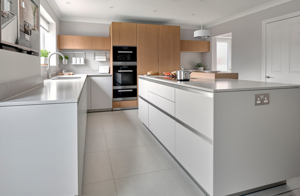 Mid-sized modern l-shaped eat-in kitchen in Oxfordshire with an integrated sink, flat-panel cabinets, grey cabinets, quartz benchtops, grey splashback, engineered quartz splashback, black appliances, porcelain floors, with island, grey floor and grey benchtop.