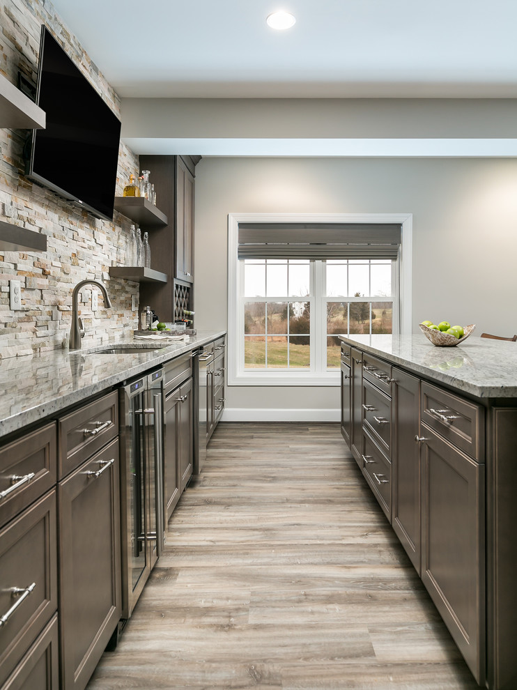 This is an example of a large contemporary single-wall wet bar in DC Metro with a drop-in sink, recessed-panel cabinets, brown cabinets, granite benchtops, beige splashback, stone tile splashback, laminate floors and brown floor.
