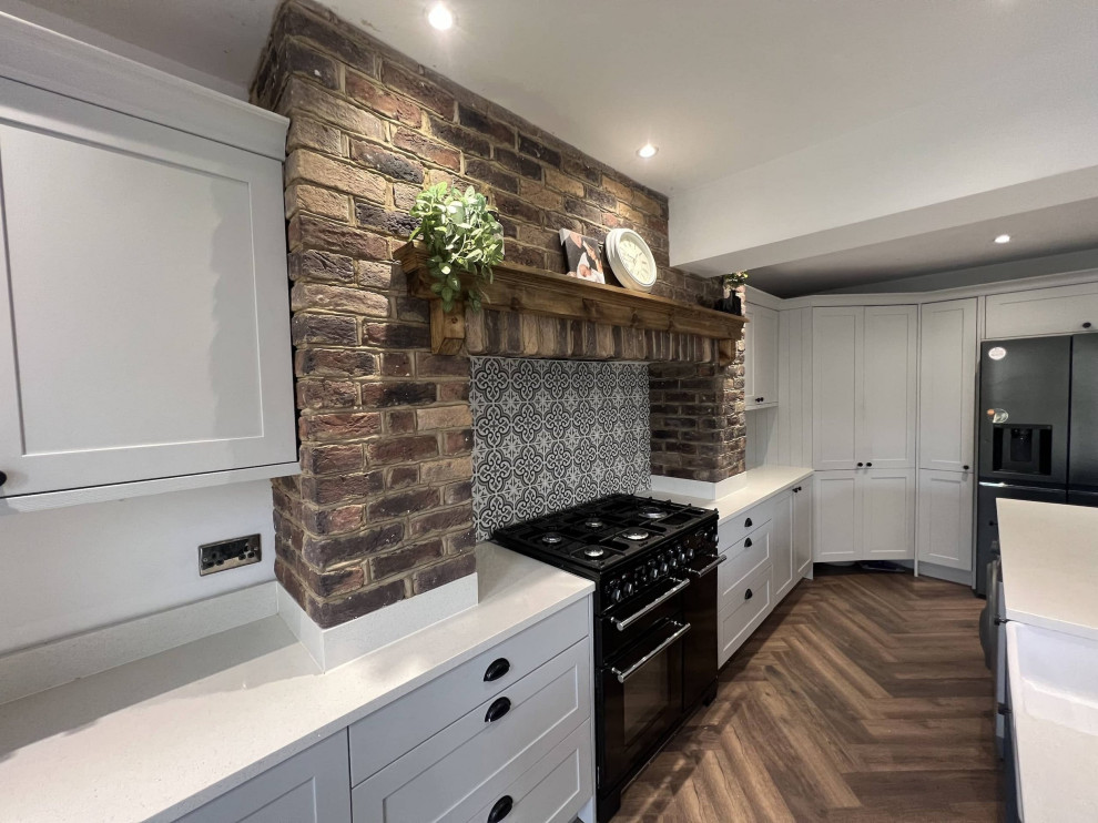 Example of a mid-sized transitional l-shaped dark wood floor and brown floor open concept kitchen design in Surrey with a farmhouse sink, recessed-panel cabinets, white cabinets, quartzite countertops, black appliances, an island and white countertops