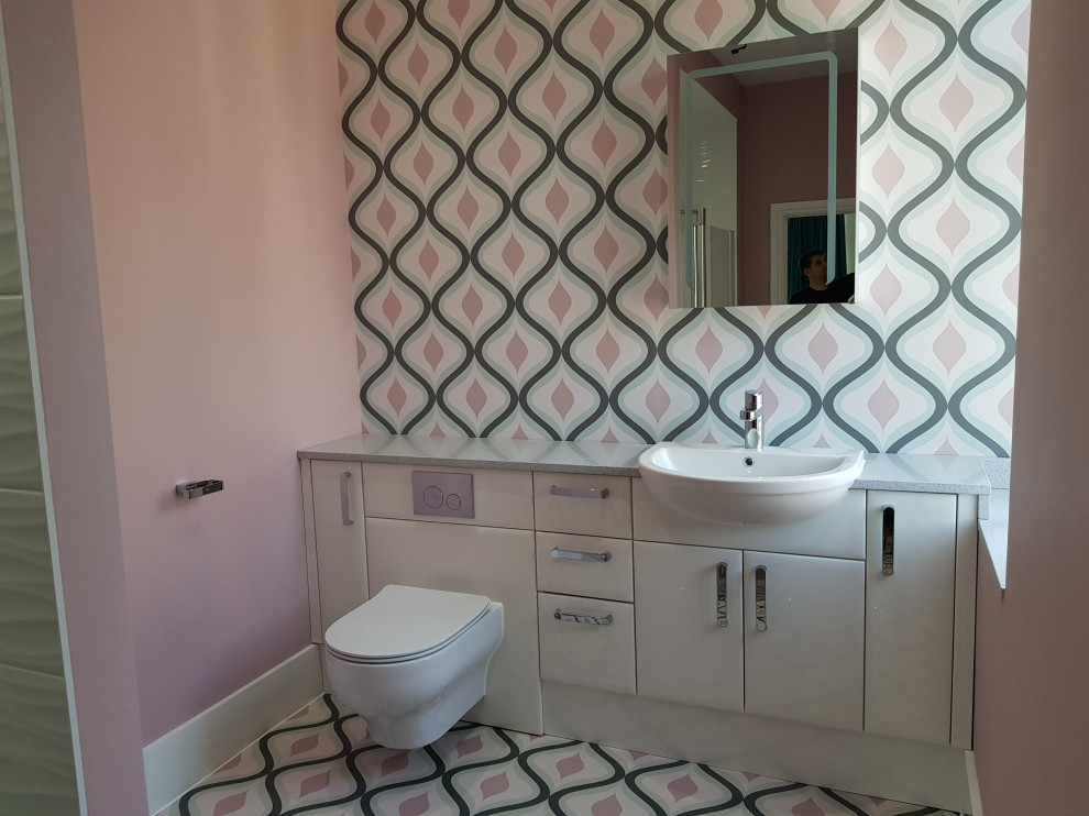 Large contemporary master bathroom in London with flat-panel cabinets, white cabinets, an open shower, a wall-mount toilet, white tile, ceramic tile, pink walls, vinyl floors, solid surface benchtops, multi-coloured floor, an open shower, grey benchtops, a single vanity, a built-in vanity and wallpaper.