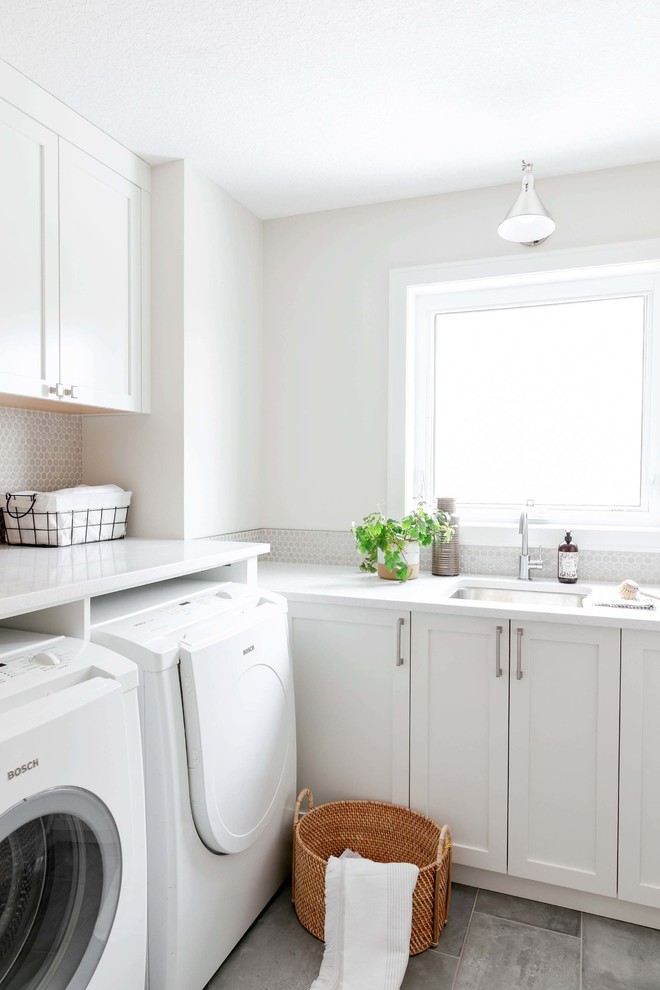Example of a mid-sized transitional l-shaped porcelain tile and gray floor utility room design in Calgary with an undermount sink, shaker cabinets, white cabinets, quartzite countertops, white walls, a side-by-side washer/dryer and white countertops