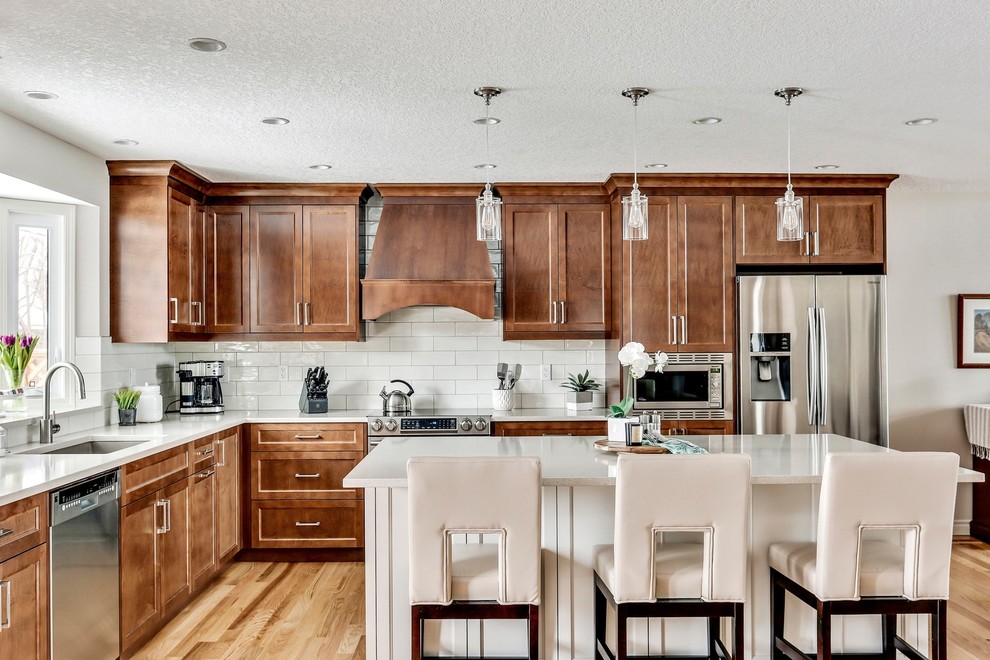 Mid-sized traditional l-shaped eat-in kitchen in Calgary with an undermount sink, shaker cabinets, dark wood cabinets, white splashback, subway tile splashback, stainless steel appliances, light hardwood floors, with island, quartzite benchtops, brown floor and white benchtop.