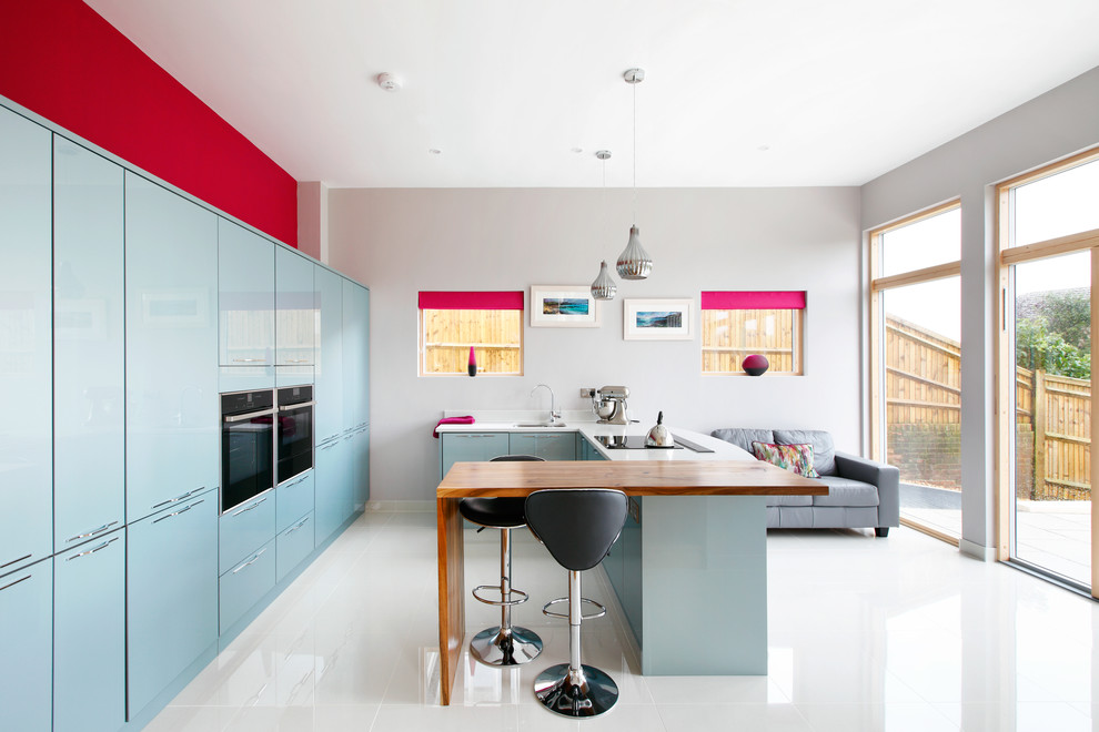 This is an example of a large contemporary u-shaped open plan kitchen in Sussex with flat-panel cabinets, blue cabinets, a drop-in sink, wood benchtops, white splashback, panelled appliances, ceramic floors and a peninsula.