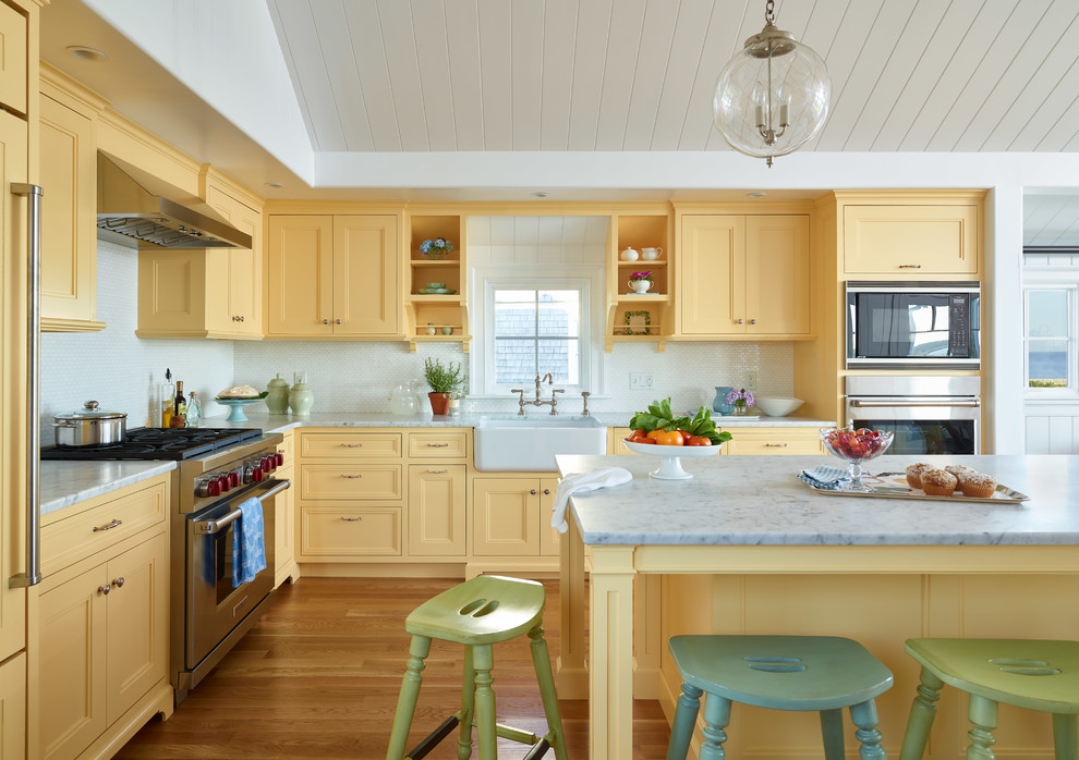 Inredning av ett maritimt vit vitt kök och matrum, med en rustik diskho, gula skåp, marmorbänkskiva, vitt stänkskydd, stänkskydd i stenkakel, integrerade vitvaror, ljust trägolv, en köksö och luckor med infälld panel