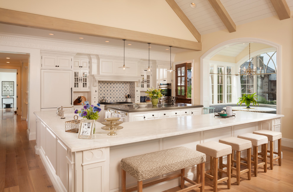 Photo of an expansive traditional l-shaped open plan kitchen in Salt Lake City with recessed-panel cabinets, white cabinets, marble benchtops, multi-coloured splashback, panelled appliances, light hardwood floors, multiple islands and brown floor.
