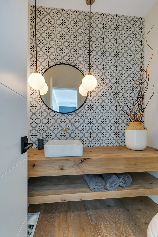 Photo of a small country powder room in Minneapolis with open cabinets, medium wood cabinets, a two-piece toilet, gray tile, ceramic tile, grey walls, medium hardwood floors, a vessel sink, wood benchtops, brown floor and brown benchtops.