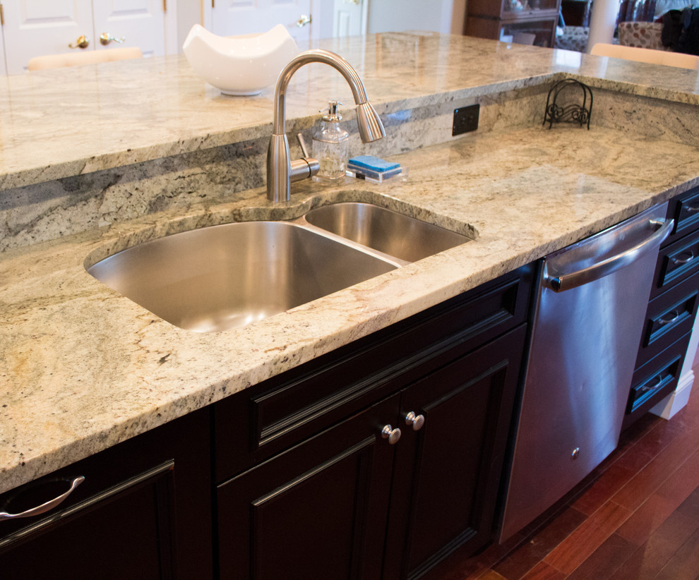 Example of a large transitional l-shaped dark wood floor and brown floor enclosed kitchen design in DC Metro with a double-bowl sink, raised-panel cabinets, dark wood cabinets, granite countertops, beige backsplash, limestone backsplash, stainless steel appliances and an island